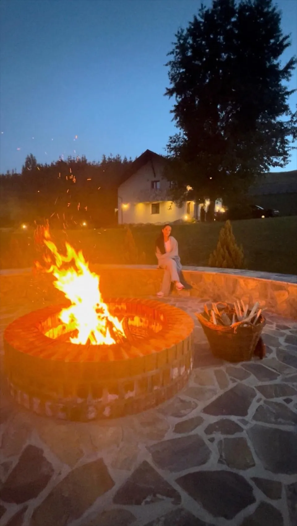 Vatra de foc aprinsă la lăsarea serii, cu scântei și vila luminată în fundal